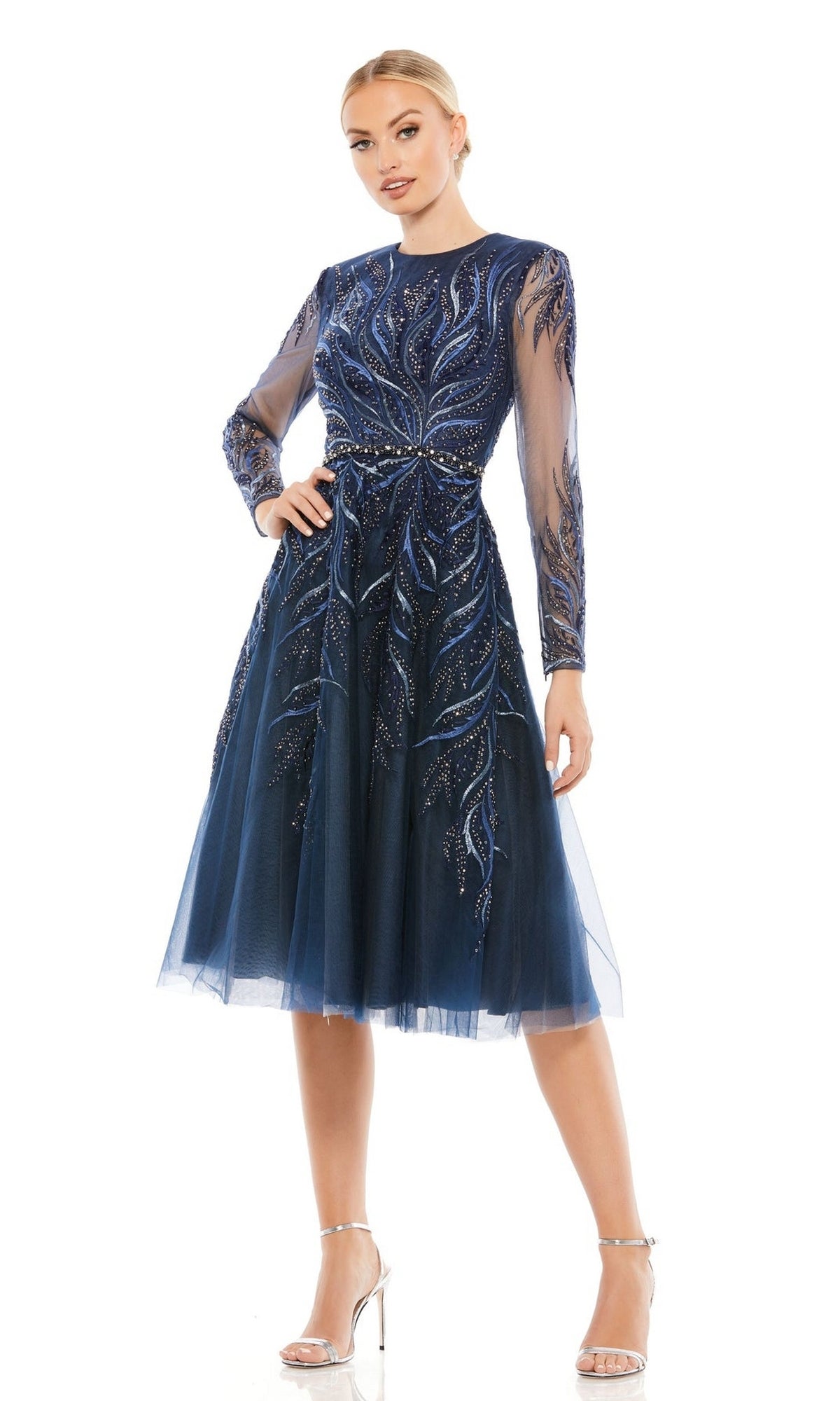Woman wearing the Mac Duggal 11136 Blue Knee-Length Cocktail Dress with sheer long sleeves, intricate beadwork, and silver high-heeled sandals, standing against a white background.